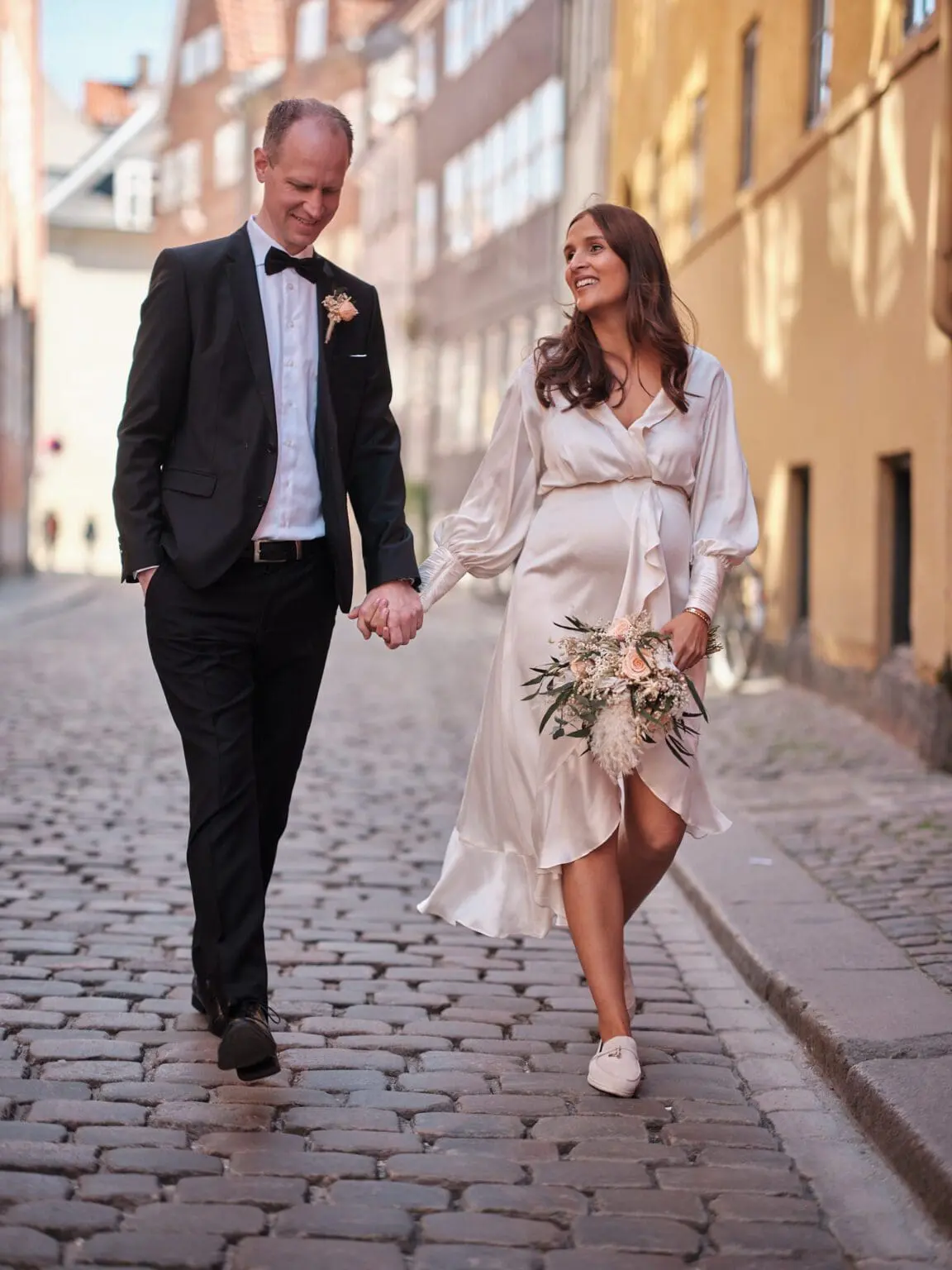 Joyful bride and groom walking hand in hand along the cobblestone street of Magstræde in Copenhagen.